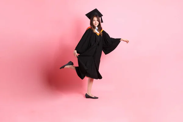 The Asian woman in graduation gown standing on the pink background.