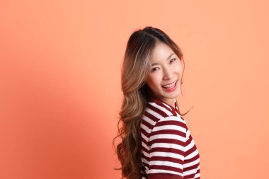 Turuncu arka planda duran ve poz veren sıradan giyinmiş genç Asyalı kadın. 