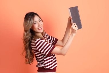 Portakal rengi arka planda kitap tutan sıradan giysili genç Asyalı kadın. 