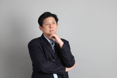 Resmi giyinmiş Asyalı iş adamı gri arka planda düşünce jesti yapıyor..