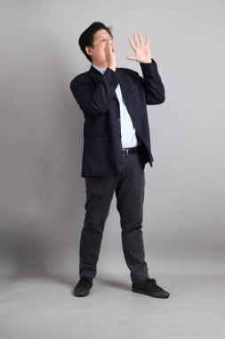 Gri arka planda bağırma hareketleriyle resmi giyinmiş Asyalı iş adamı..