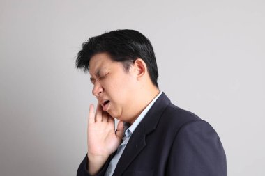 Gri arka planda diş ağrısı jesti ile resmi giyinmiş Asyalı iş adamı..