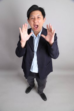 Gri arka planda bağırma hareketleriyle resmi giyinmiş Asyalı iş adamı..