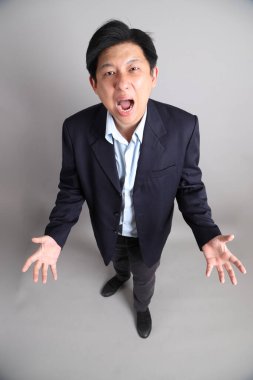 Asyalı iş adamı. Resmi giyinmiş. Gri arka planda kızgın ve çığlık çığlığa bir jestle..