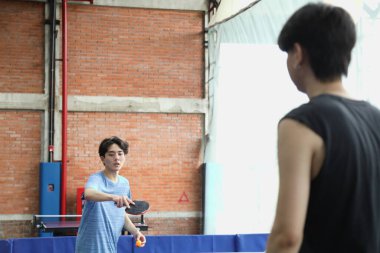 Arkadaşı olan Asyalı yakışıklı bir genç adam elinde bir top ve raket tutarken masa tenisi kulübünde masa tenisi oynuyor.