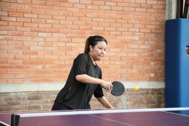 Asyalı genç bir kadın halka açık masa tenisi kulübünde elinde bir top ve raket tutarken masa tenisi oynuyor.