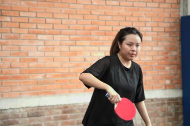 Asyalı genç bir kadın halka açık masa tenisi kulübünde elinde bir top ve raket tutarken masa tenisi oynuyor.