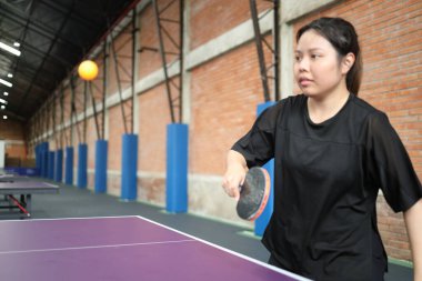 Asyalı genç bir kadın halka açık masa tenisi kulübünde elinde bir top ve raket tutarken masa tenisi oynuyor.