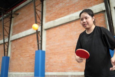 Asyalı genç bir kadın halka açık masa tenisi kulübünde elinde bir top ve raket tutarken masa tenisi oynuyor.