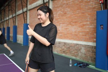 Asya 'lı genç bir kadın halka açık masa tenisi kulübünde elinde bir top ve raket tutarken gülümseyerek ping pong oynuyor.