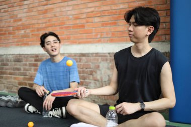 Asyalı arkadaşlar halka açık masa tenisi kulübünde elinde bir top ve raket tutarken birlikte masa tenisi oynuyorlar.