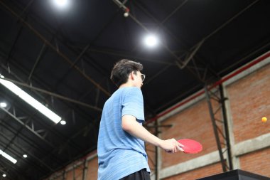Asyalı yakışıklı genç adam halka açık masa tenisi kulübünde elinde bir top ve raket tutarken masa tenisi oynuyor.