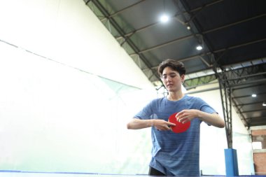 Asyalı yakışıklı genç adam halka açık masa tenisi kulübünde elinde bir top ve raket tutarken masa tenisi oynuyor.