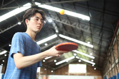 Asyalı yakışıklı genç adam halka açık masa tenisi kulübünde elinde bir top ve raket tutarken masa tenisi oynuyor.