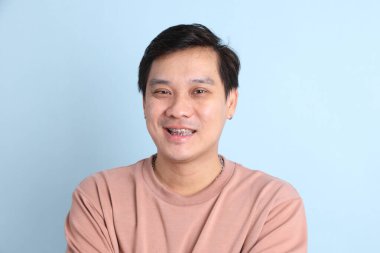 Diş teli takan, sıradan bir tişört giyen, mavi arka planda gülümseyen Asyalı bir adam..