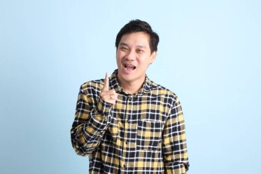 Diş teli takan, sıradan bir tişört giyen, mavi arka planda düşünme hareketi yapan Asyalı bir adam..