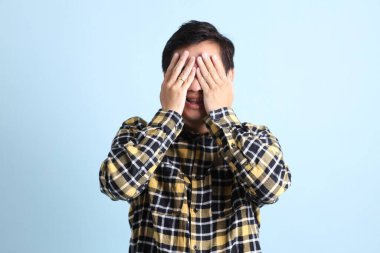 Diş teli takan Asyalı bir adam, sıradan bir tişört giyiyor, mavi arka planda gizli bir göz hareketi yapıyor..