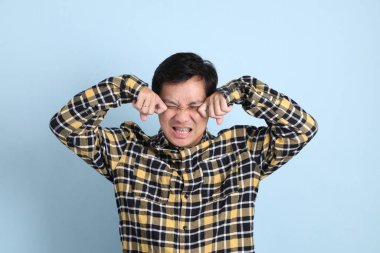 Asyalı bir adam, sıradan bir tişört giyiyor, hayal kırıklığı jesti, mavi arka planda ağlıyor..