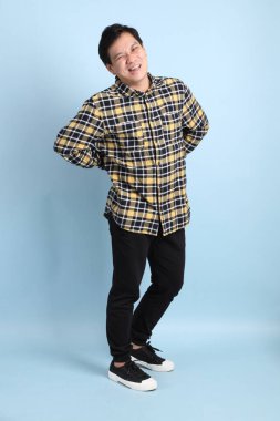 Diş teli takan Asyalı bir adam, günlük tişört giyiyor, mavi arka planda sırt ağrısı jesti yapıyor..