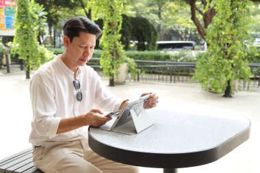 40 yaşındaki Asyalı işadamı gündüz vakti açık bir yerde tablet üzerinde çalışıyor..