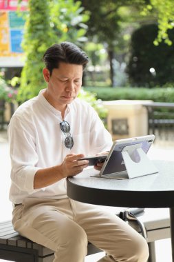 40 yaşındaki Asyalı işadamı gündüz vakti açık bir yerde tablet üzerinde çalışıyor..