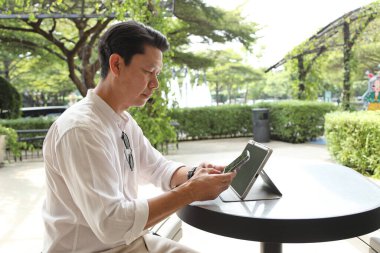 40 yaşındaki Asyalı işadamı gündüz vakti açık bir yerde tablet üzerinde çalışıyor..