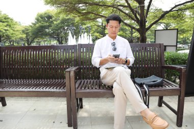 40 yaşındaki Asyalı işadamı bankta oturuyor, akıllı telefon kullanıyor..