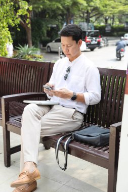 40 yaşındaki Asyalı işadamı bankta oturuyor, akıllı telefon kullanıyor..