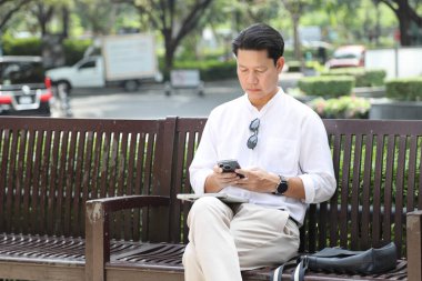 40 yaşındaki Asyalı işadamı bankta oturuyor, akıllı telefon kullanıyor..