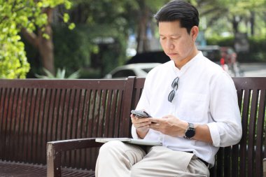 40 yaşındaki Asyalı işadamı bankta oturuyor, akıllı telefon kullanıyor..