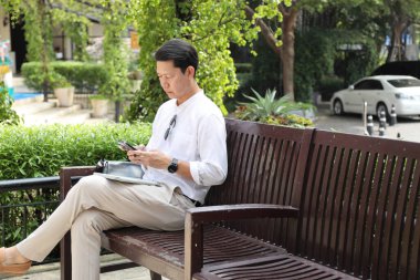 40 yaşındaki Asyalı işadamı bankta oturuyor, akıllı telefon kullanıyor..