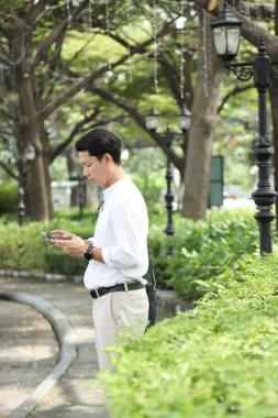 40 yaşındaki Asyalı işadamı park yerinde gündüz vakti akıllı telefon kullanarak yürüyor..