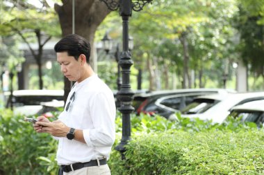 40 yaşındaki Asyalı işadamı park yerinde gündüz vakti akıllı telefon kullanarak yürüyor..
