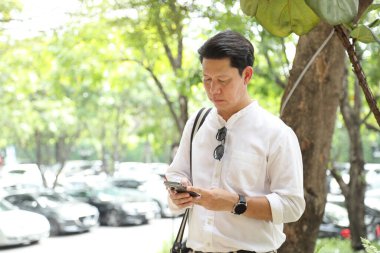 40 yaşındaki Asyalı işadamı park yerinde gündüz vakti akıllı telefon kullanarak yürüyor..