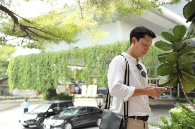 40 yaşındaki Asyalı işadamı park yerinde gündüz vakti akıllı telefon kullanarak yürüyor..