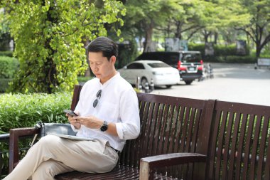 40 yaşındaki Asyalı işadamı bankta oturuyor, akıllı telefon kullanıyor..