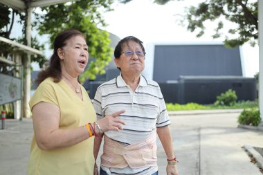 Bel destek kemeri takan kıdemli Asyalı adam gün içinde karısıyla parkta yürüyor..