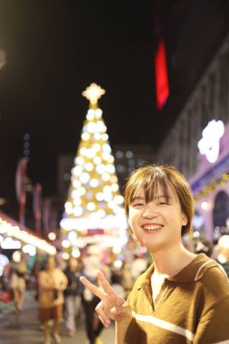 Doğu Asyalı kadın kameraya gülümsüyor, Noel arifesinde arka planda Noel etkinliğinden kalma bokeh ışıkları var..