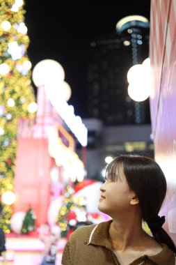 Doğu Asyalı kadın kameraya gülümsüyor, Noel arifesinde arka planda Noel etkinliğinden kalma bokeh ışıkları var..