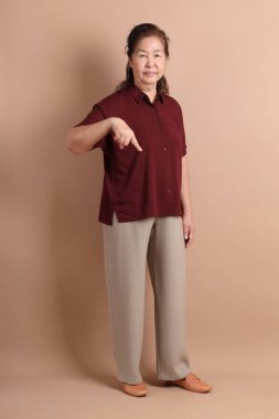 Kahverengi arka planda duran koyu kırmızı tişörtlü kıdemli Doğu Asyalı kadın..