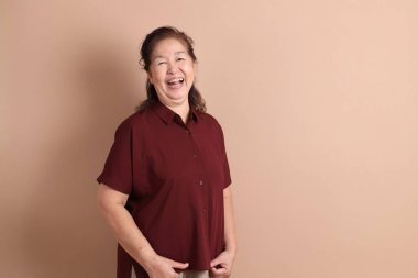 Kahverengi arka planda duran koyu kırmızı tişörtlü kıdemli Doğu Asyalı kadın..