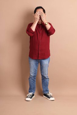 Kahverengi arka planda duran koyu kırmızı gömlekli kıdemli Doğu Asyalı adam..