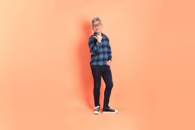 Koyu mavi ekose gömlekli yaşlı bir Asyalı şeftali pembesi bir arka plana karşı duruyor..