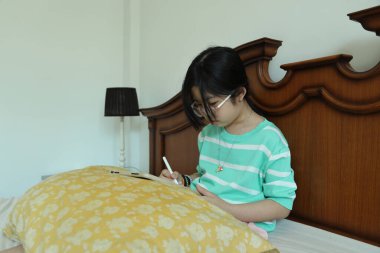 Doğu Asyalı kız yatakta otururken dijital tablet üzerinde ödev yapıyor..