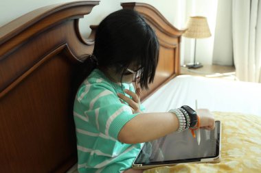 Doğu Asyalı kız yatakta otururken dijital tablet üzerinde ödev yapıyor..