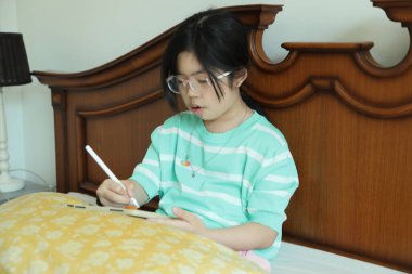 Doğu Asyalı kız yatakta otururken dijital tablet üzerinde ödev yapıyor..