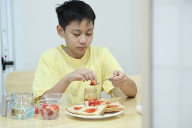 Güneydoğu Asyalı çocuk çilekli sandviç yapıyor ve evde tek başına yiyor..