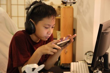 Güneydoğu Asyalı çocuk bilgisayar masasında akıllı telefonuyla oyun oynuyor..