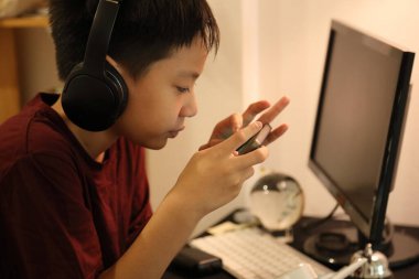 Güneydoğu Asyalı çocuk bilgisayar masasında akıllı telefonuyla oyun oynuyor..
