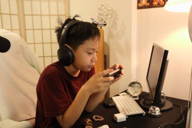 Güneydoğu Asyalı çocuk bilgisayar masasında akıllı telefonuyla oyun oynuyor..